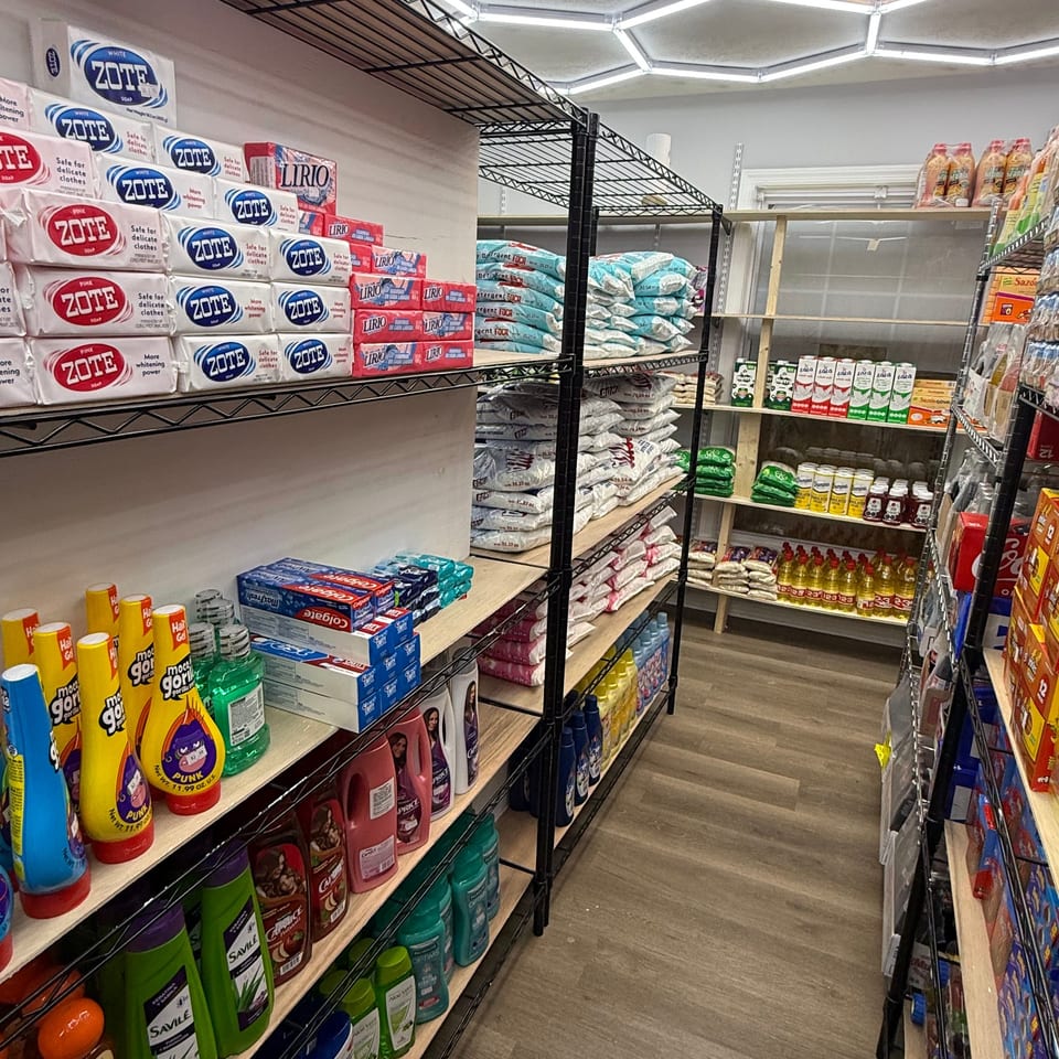 Grocery store aisle stocked with detergent, cleaning products, rice, cooking oil, and packaged household goods on metal shelves.